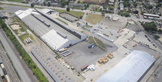 Aerial view of the Modu Tech industrial facility, showcasing large warehouse buildings, outdoor material storage yards, and rail access.